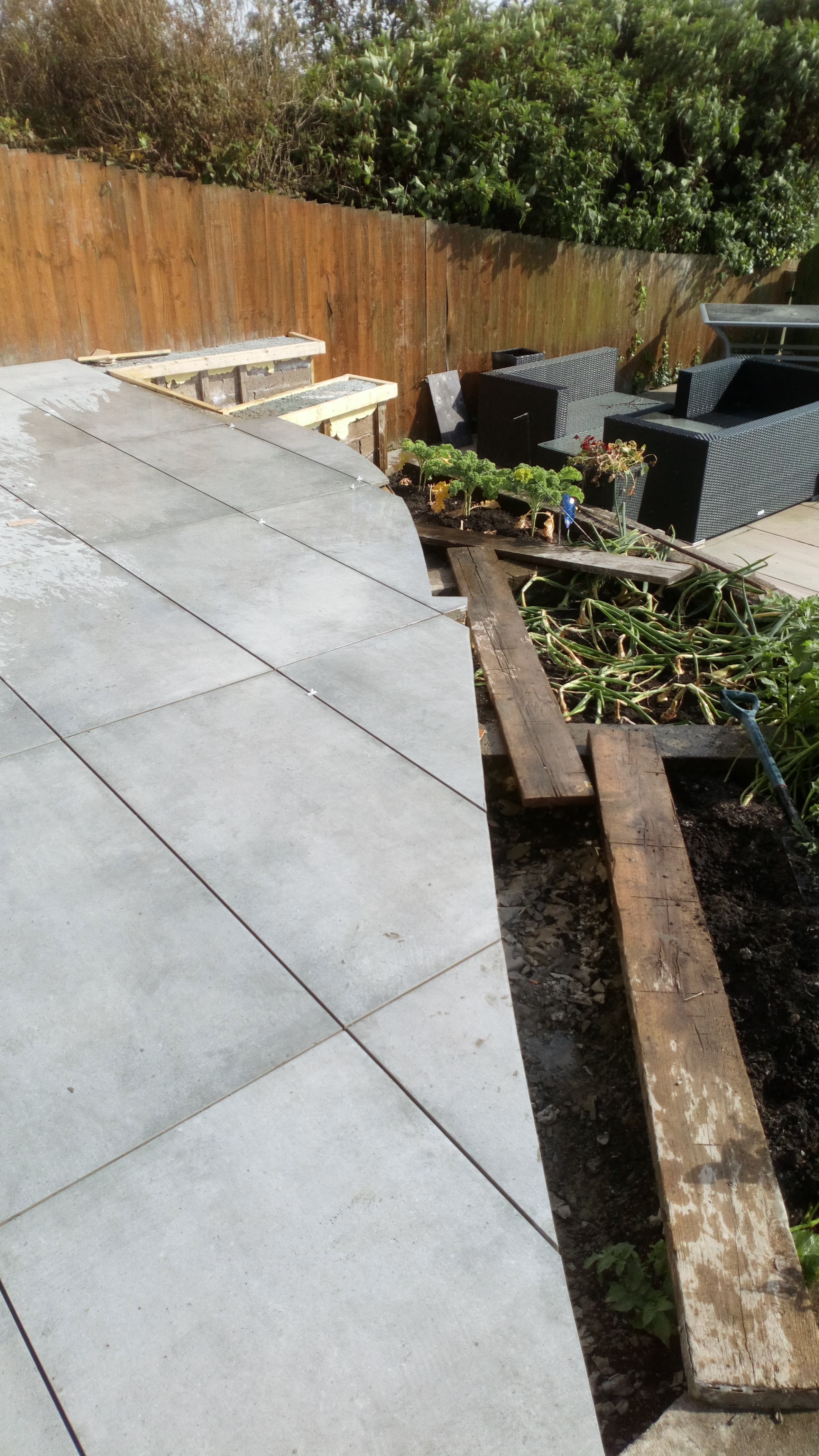 Curved porcelain patio mid-build, wider view with railway-sleeper raised beds in shot