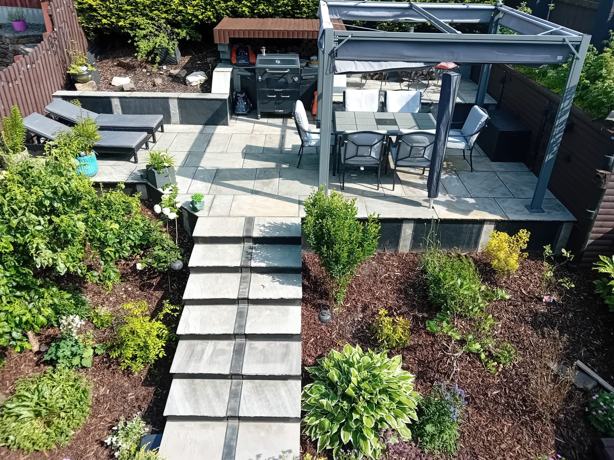 Tiered back garden seen from above in daylight — upper flagged terrace with pergola and outdoor kitchen, lower lounge terrace, stepped sandstone path running between, planted beds either side