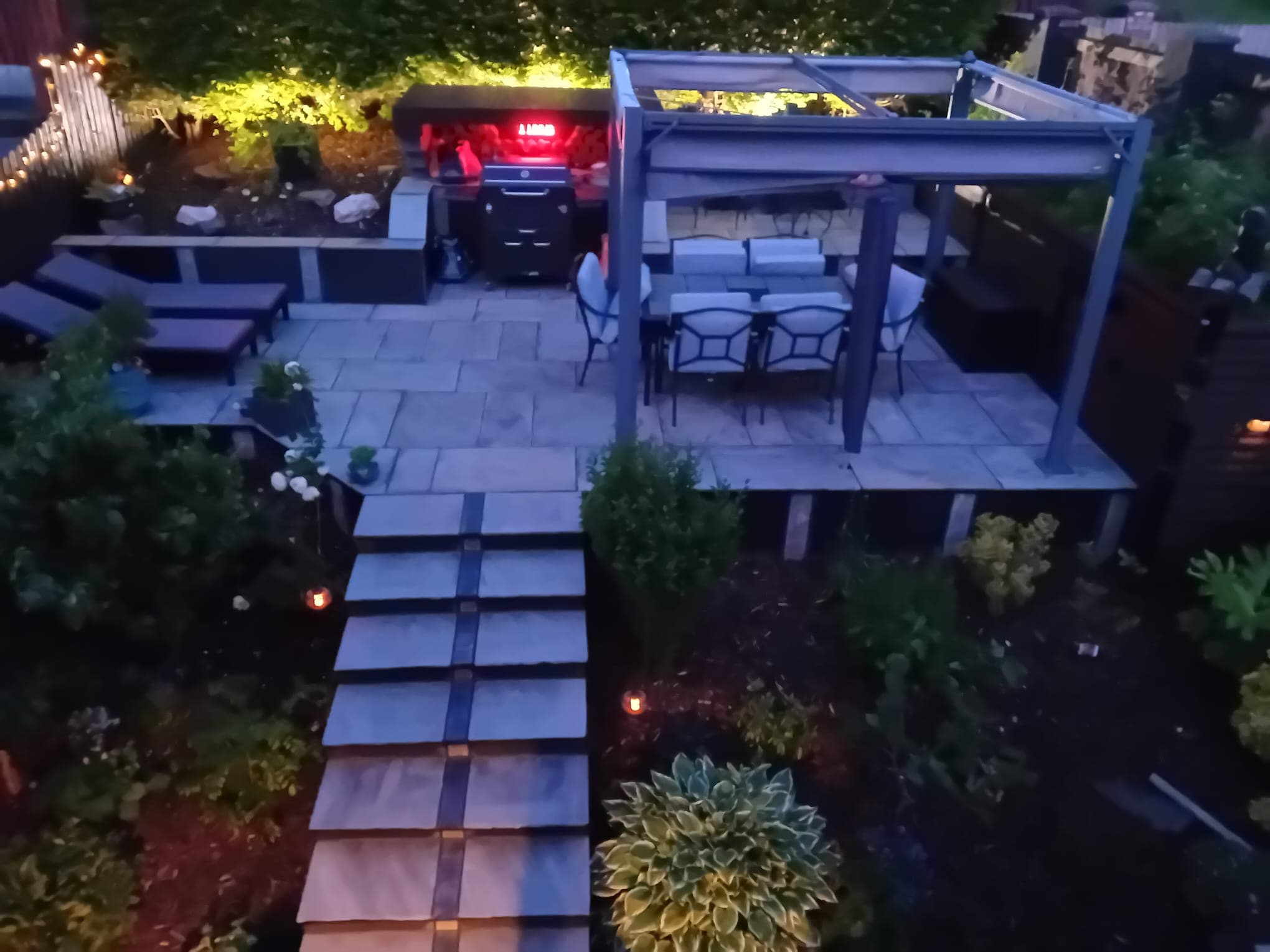 Same tiered garden at dusk — upper terrace lit, BBQ glowing inside the pergola, dining table set, lounge terrace and planting lit by garden lights below
